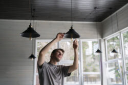 A man changing a light bulb in a hanging ceiling light fixture, with natural daylight illuminating the scene.